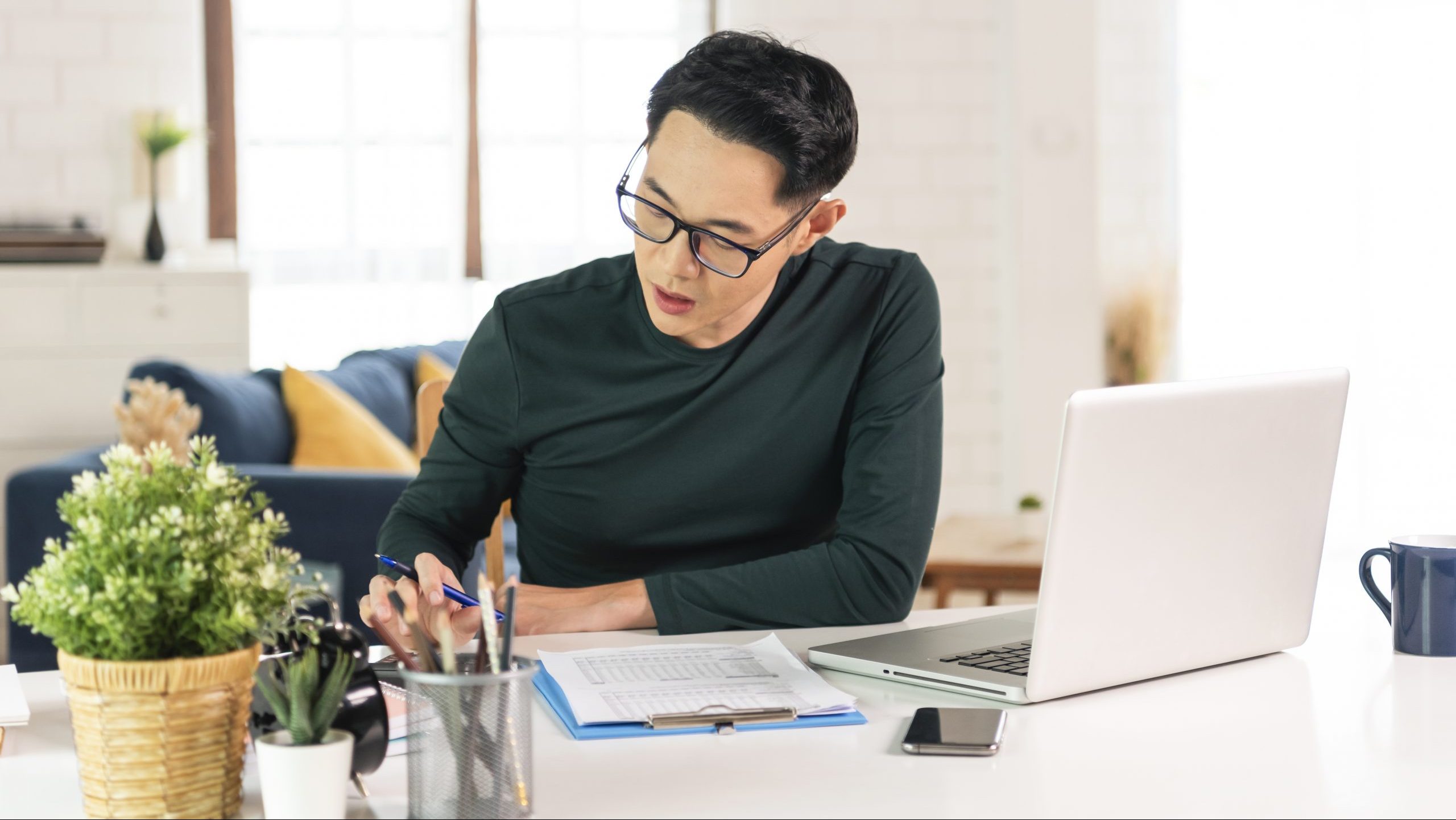 Asian man working at home with laptop and papers on desk.