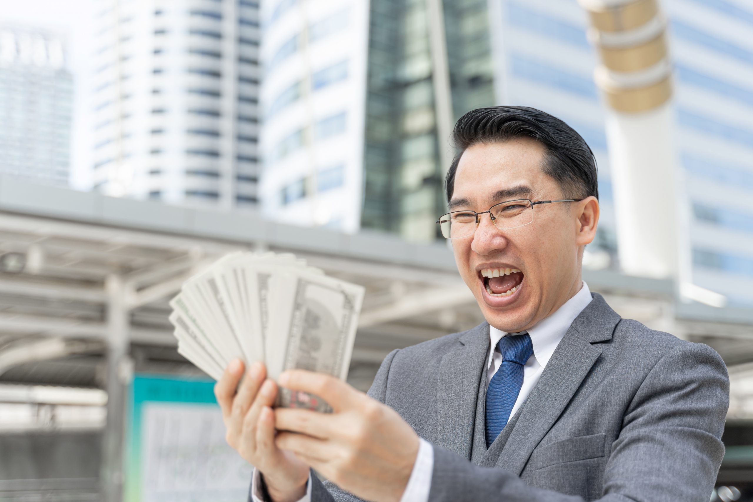 Asian man in suit happily looking at a fan of cash he's holding.