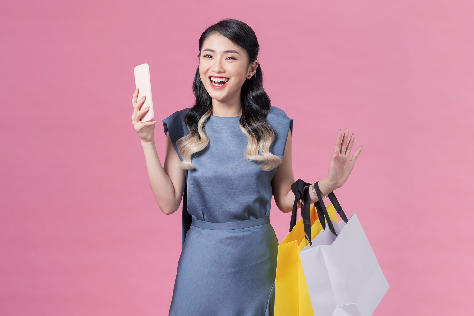 happy young woman holding shopping bags and mobile phone over pink background