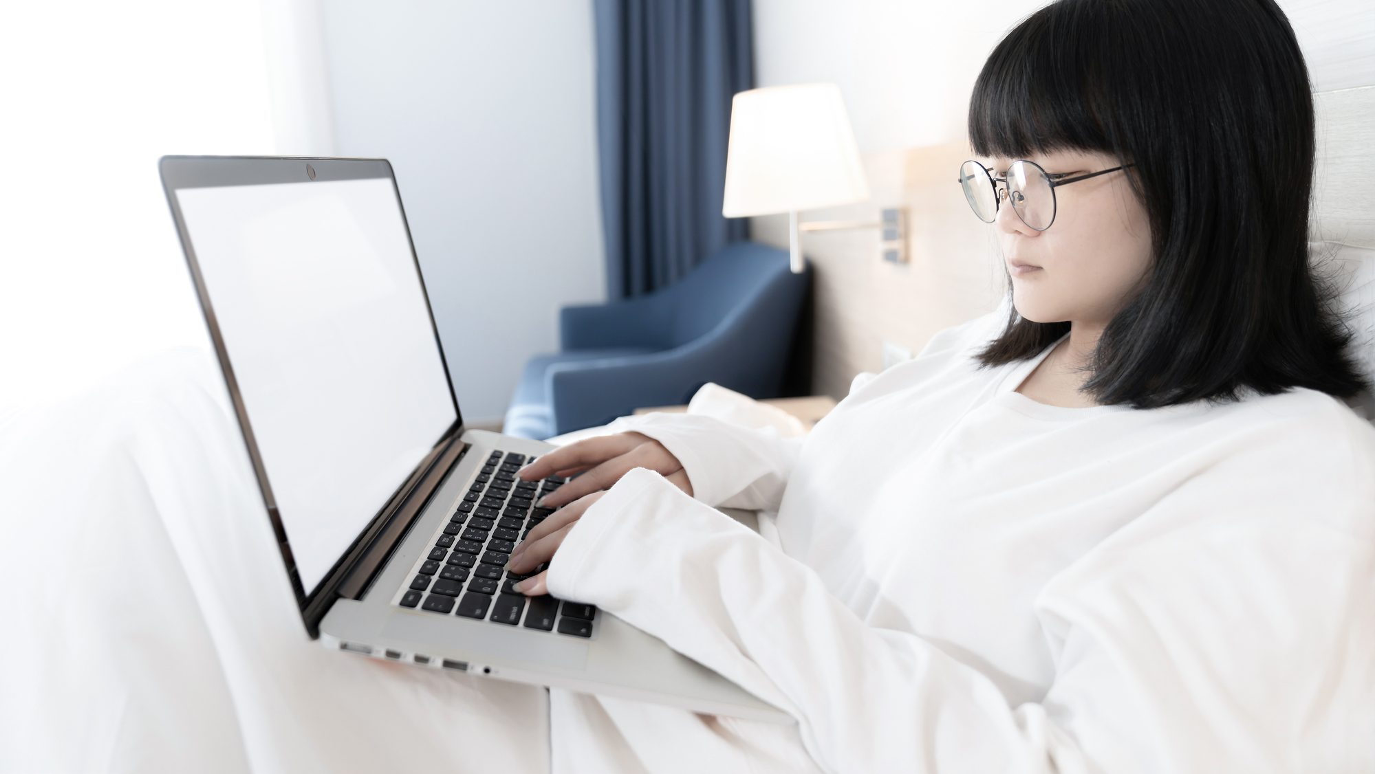 Asian woman with glasses working on her laptop in bed.