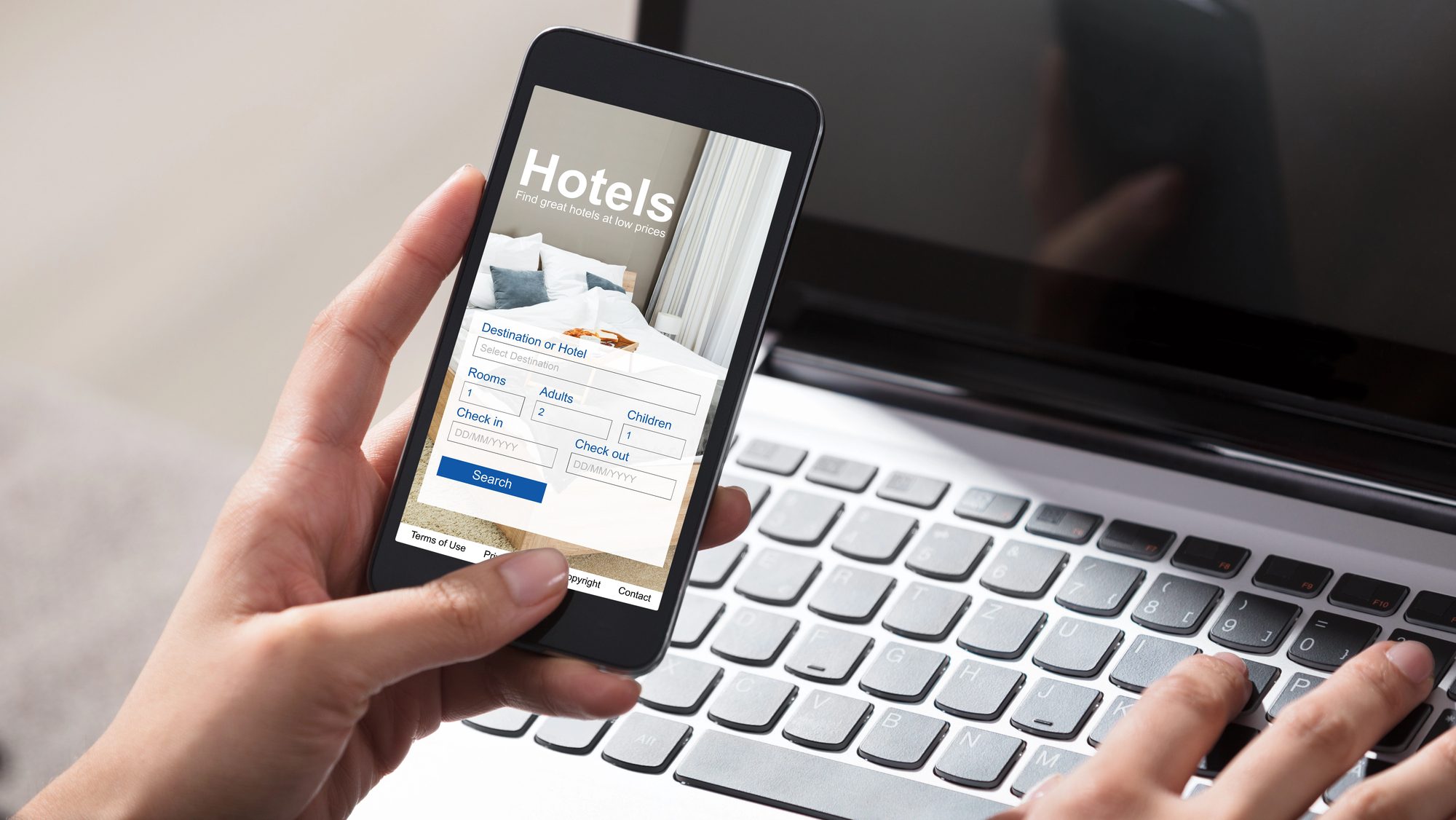 person's hand holding phone booking a hotel while typing on a laptop