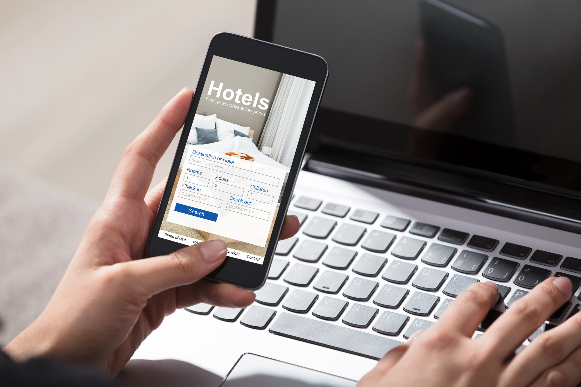 person's hand holding phone booking a hotel while typing on a laptop