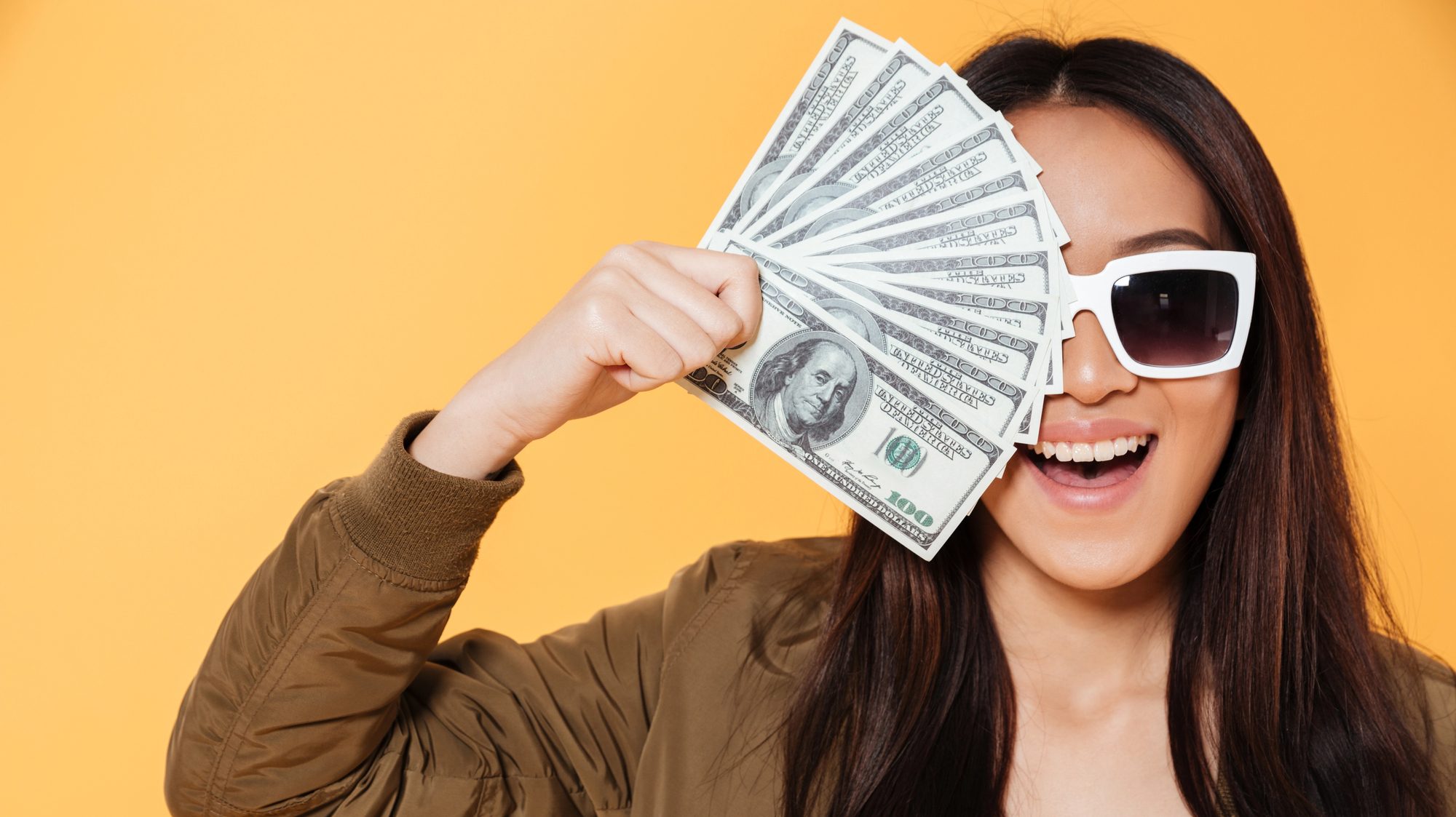 Woman wearing sunglasses covering half of her face with a fan of dollar bills.