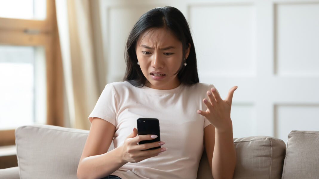 Mad Asian woman sitting on a couch while holding her smartphone.