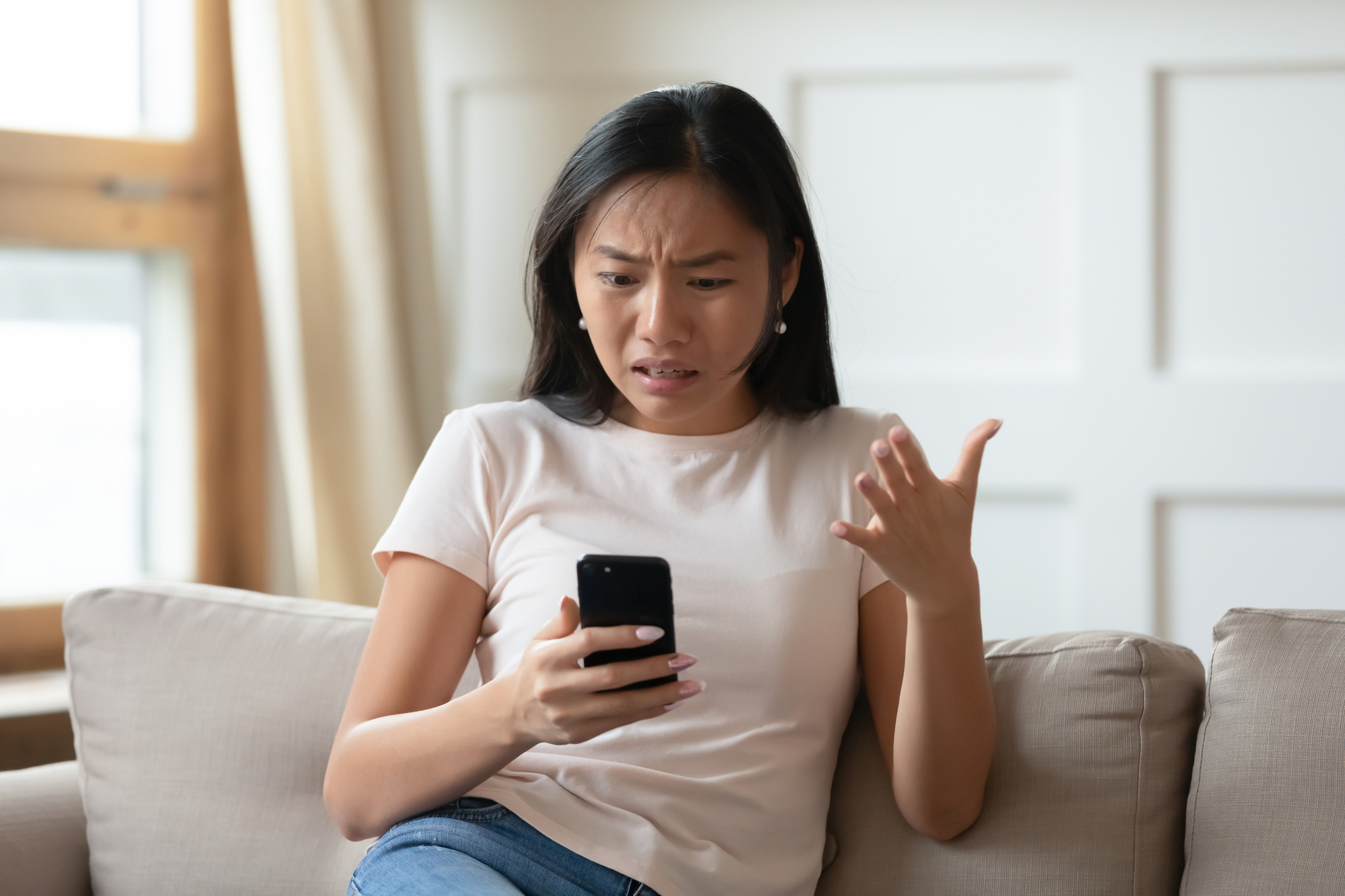 Mad Asian woman sitting on a couch while holding her smartphone.