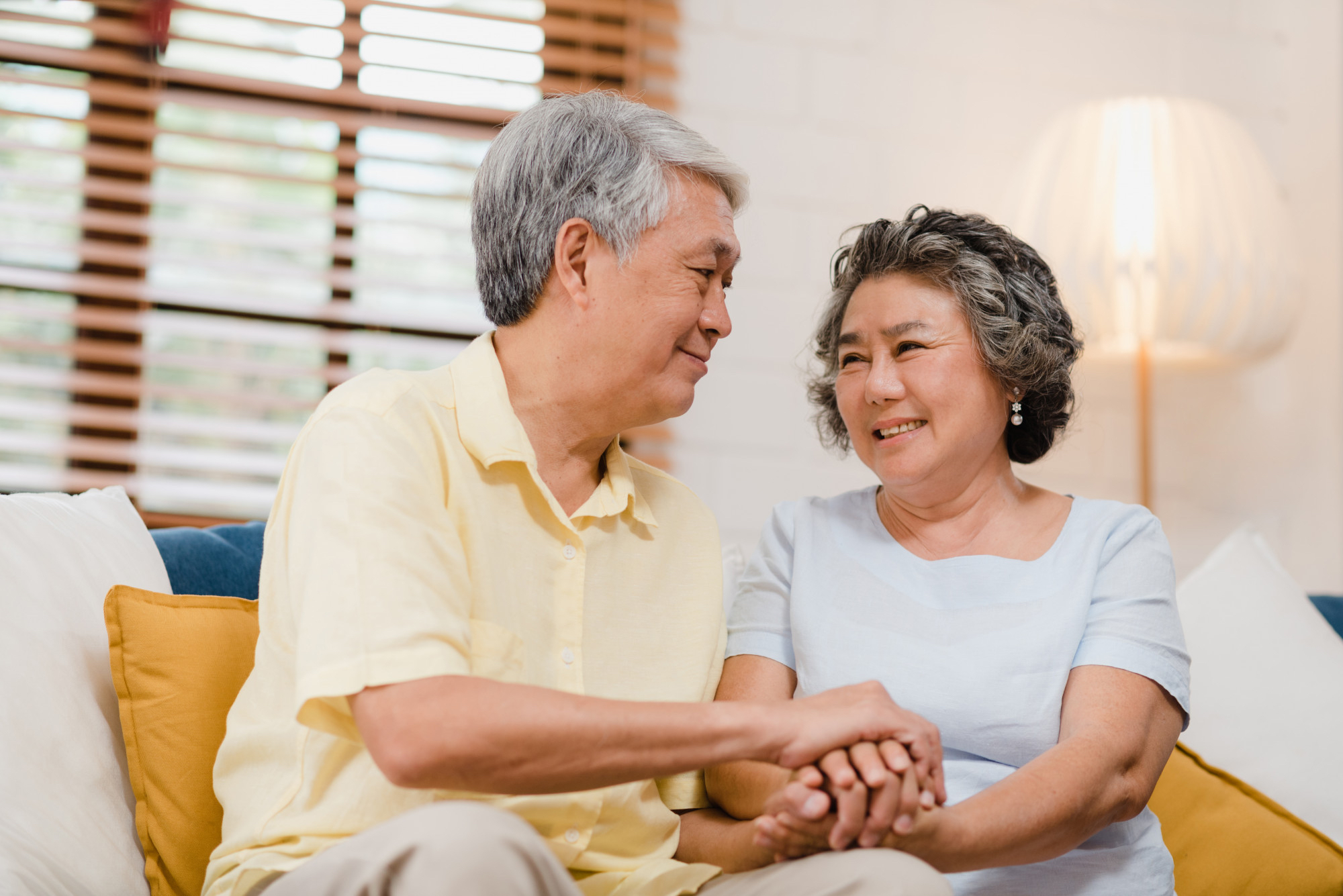 Happy Asian elderly couple holding hands while smiling and looking at each other in the living room.