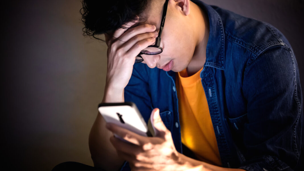 A man wearing glasses and a denim shirt looks stressed while holding his forehead and a smartphone, conveying a sense of worry or frustration