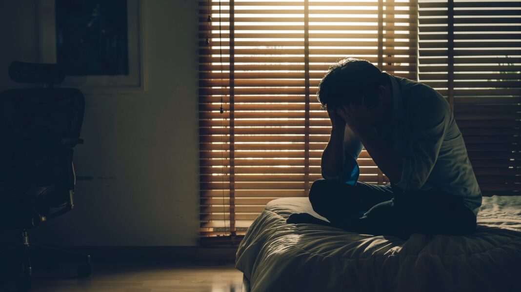 A male individual sits on a bed in a dimly lit room, head in hands, silhouetted against light streaming through window blinds, conveying a sense of stress, sadness or depression