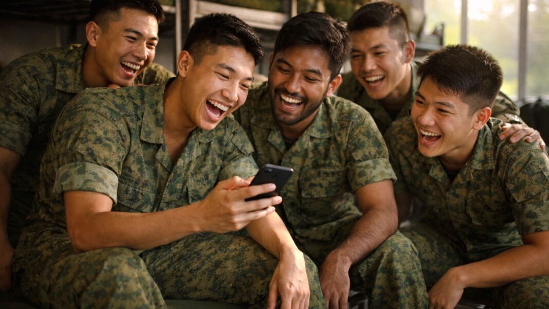 Five soldiers (NS men) in camouflage uniforms sit closely, joyfully laughing while looking at a smartphone. The scene conveys nostalgic memories, camaraderie and happiness