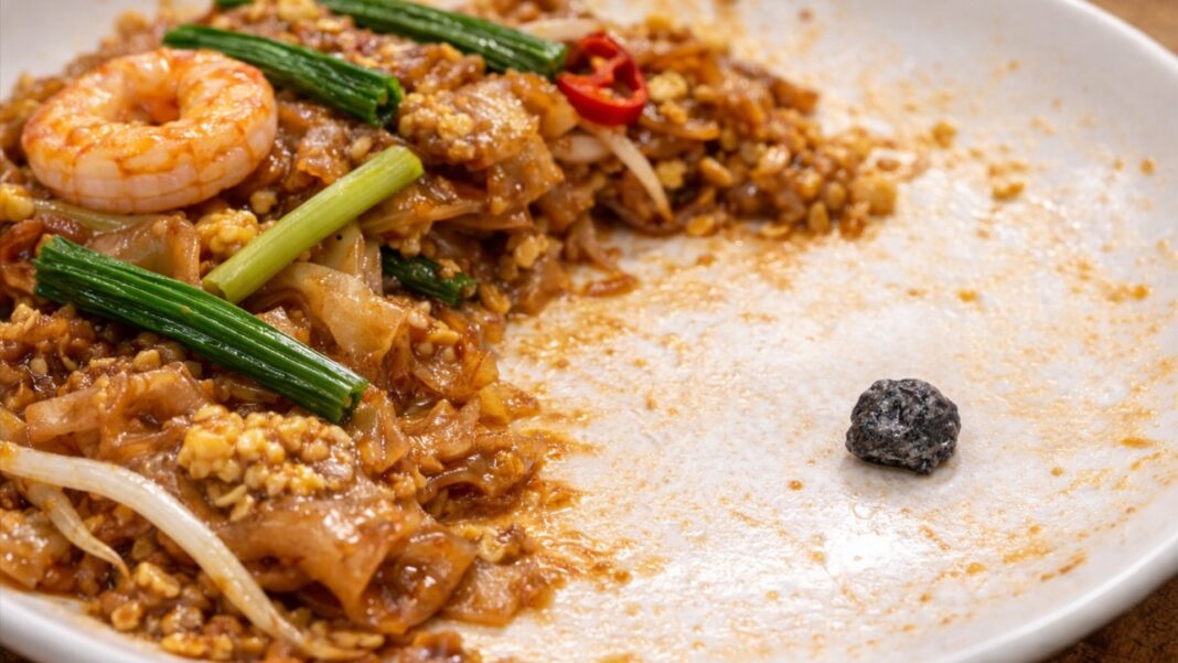 A partially eaten plate of Thai-style fried kway teow, with a small stone visible on the empty section of the plate