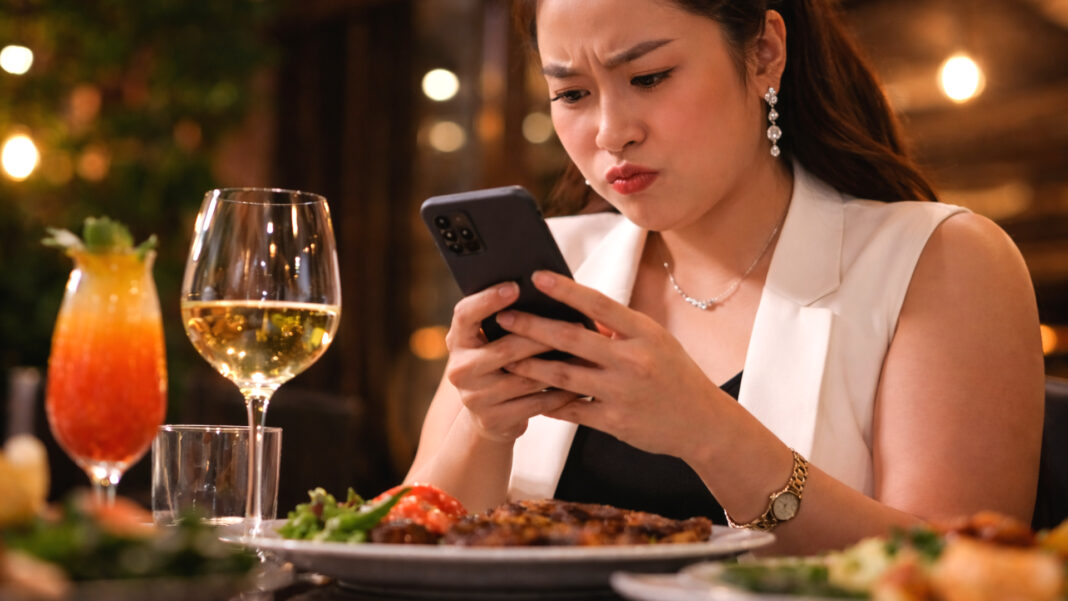 A well-dressed woman looks visibly displeased as she scrolls through her phone at a high-end dining table, where expensive food and drinks sit untouched, reflecting tensions.