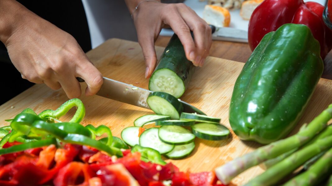 Hands slicing a cucumber on a wooden cutting board with a knife. Surrounding are green bell pepper, asparagus, and red pepper, evoking freshness