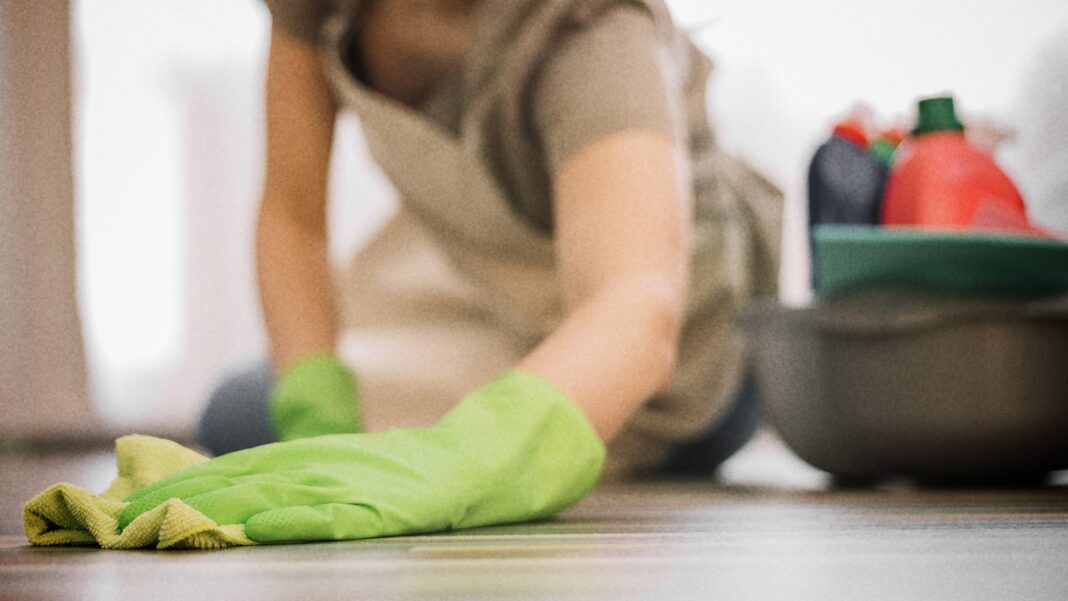 A woman in a beige apron and green gloves scrubs a wooden floor, surrounded by cleaning supplies, as the scene conveys cleaning and tidying up