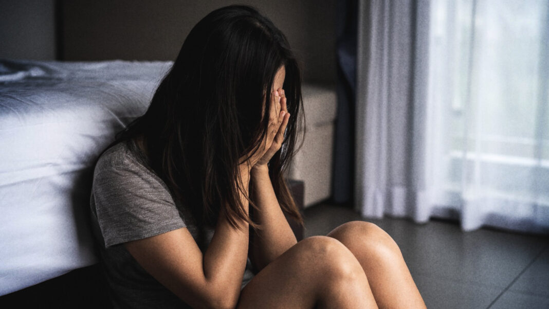 A woman in a gray shirt sits on the floor, leaning against a bed, with her face buried in her hands, while the room is dimly lit, conveying a sense of sadness and distress