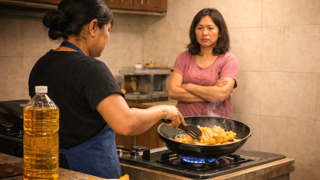 A domestic helper deep-fries food at the stove while her employer watches closely in the background, capturing the quiet strain that can build when household expectations are misaligned