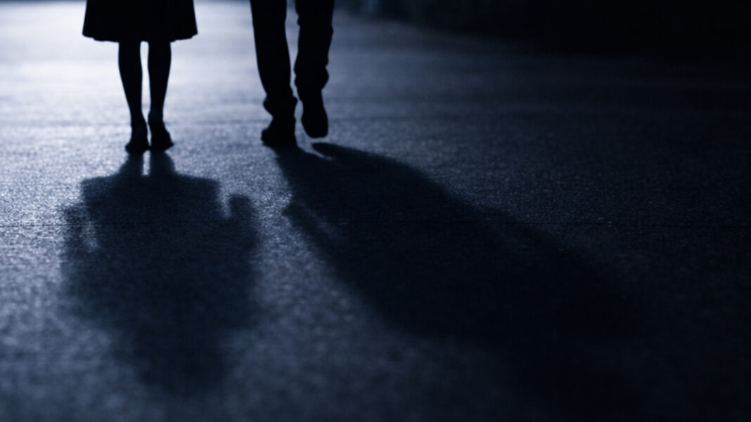 Two silhouetted figures walk side by side along a quiet, blue-lit path at night—identical in scale, indistinct in identity—leaving only their shadows and the stillness of the street behind them